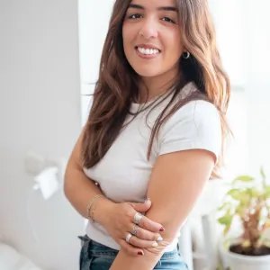 Woman smiling indoors by a window, wearing a white shirt and jeans, plants in background.