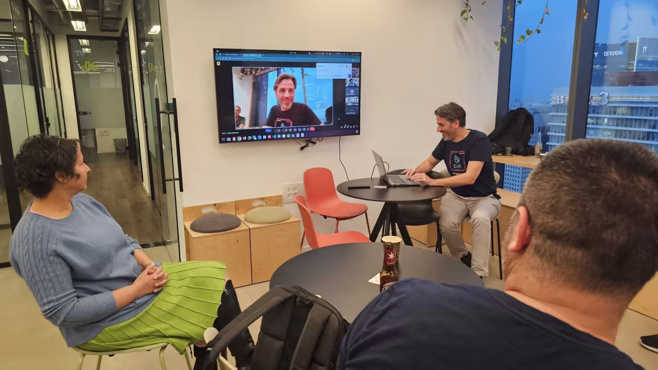 Three people sit in an office watching a video call on a wall-mounted screen.