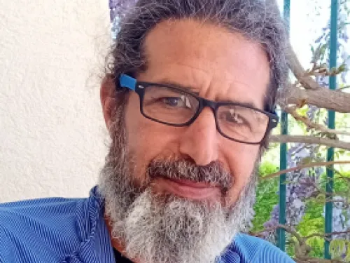 Man with glasses and gray beard smiling outdoors near a wall and some plants.