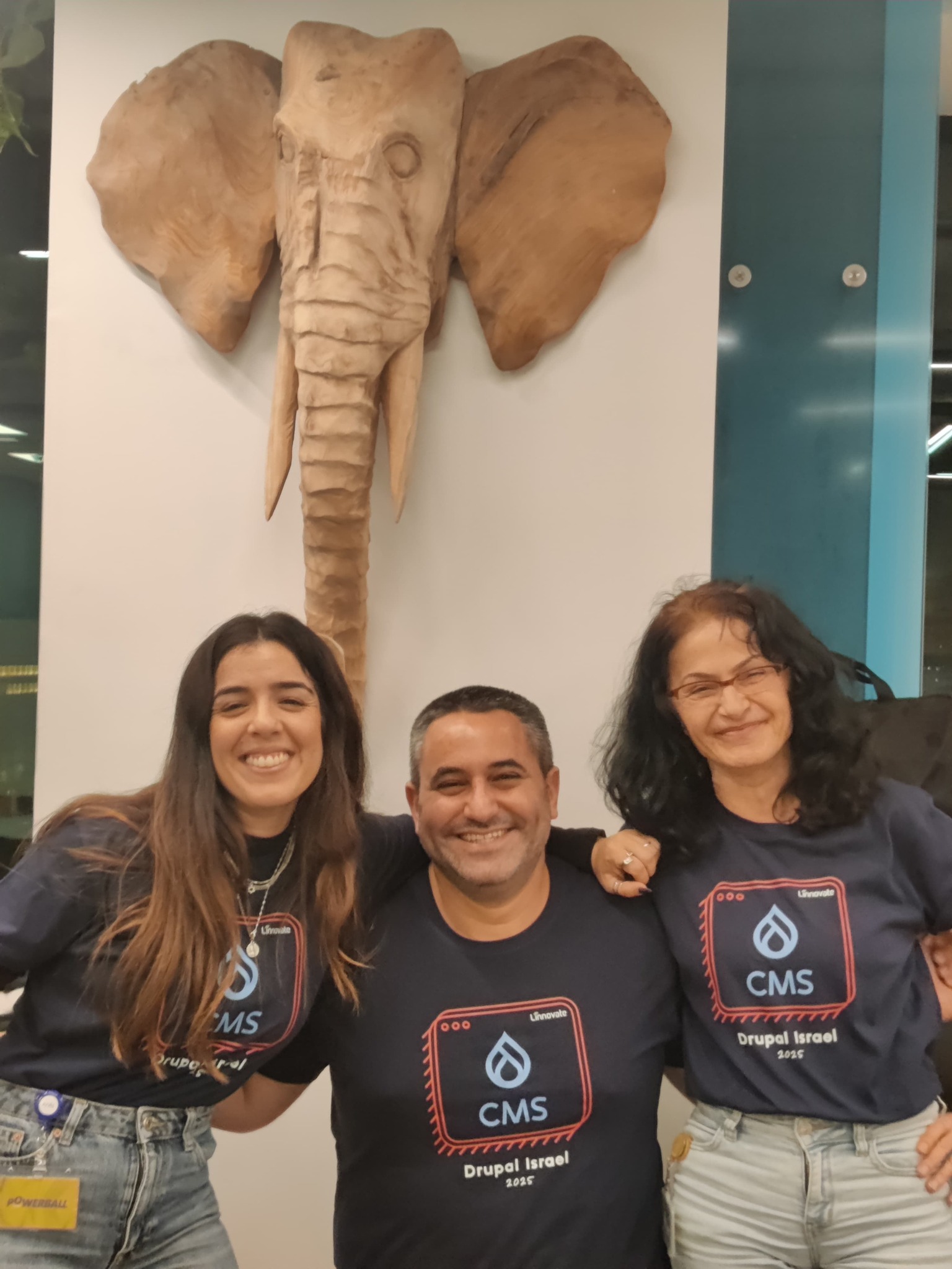Three people smiling in front of an elephant wall sculpture.