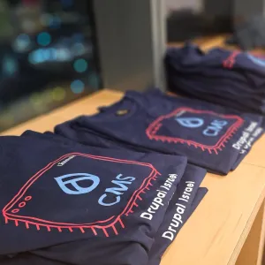 Stack of folded navy blue CMS Drupal Israel t-shirts on a wooden table.