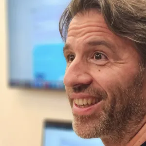 Man smiling indoors with blurred monitor and window in the background