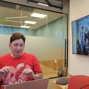 Person in red shirt at desk, group photo displayed on wall-mounted monitor.