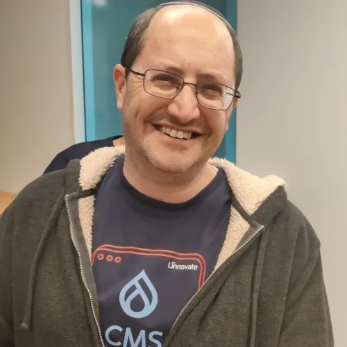 Smiling man wearing glasses, a zip-up hoodie, and a CMS t-shirt indoors.