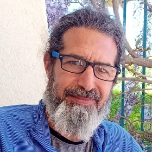 Man with glasses and gray beard smiling outdoors near a wall and some plants.