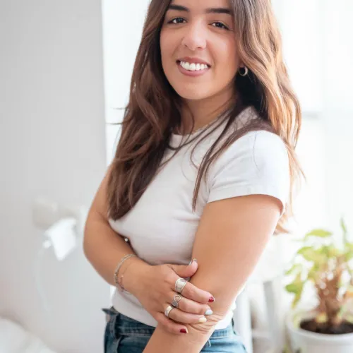Woman smiling indoors by a window, wearing a white shirt and jeans, plants in background.