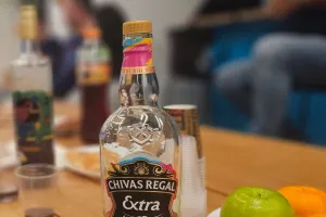 Chivas Regal whisky bottle on a table with fruit, cups, and blurred people in the background.