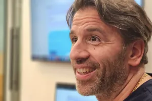 Man smiling indoors with blurred monitor and window in the background