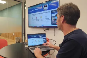Man working on a laptop with a presentation displayed on a wall-mounted screen.
