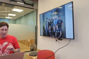Man in red shirt sitting at desk, group photo displayed on wall-mounted screen.