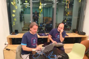 Two drupal gurus in matching shirts work on laptops at a round table by a window