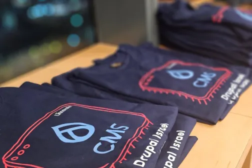 Stack of folded navy blue CMS Drupal Israel t-shirts on a wooden table.