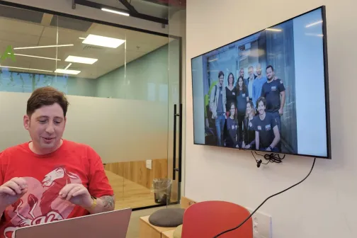 Person in red shirt at desk, group photo displayed on wall-mounted monitor.