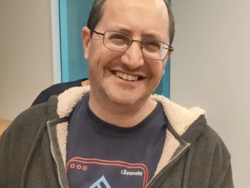 Smiling man wearing glasses, a zip-up hoodie, and a CMS t-shirt indoors.
