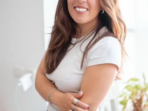Woman smiling indoors by a window, wearing a white shirt and jeans, plants in background.
